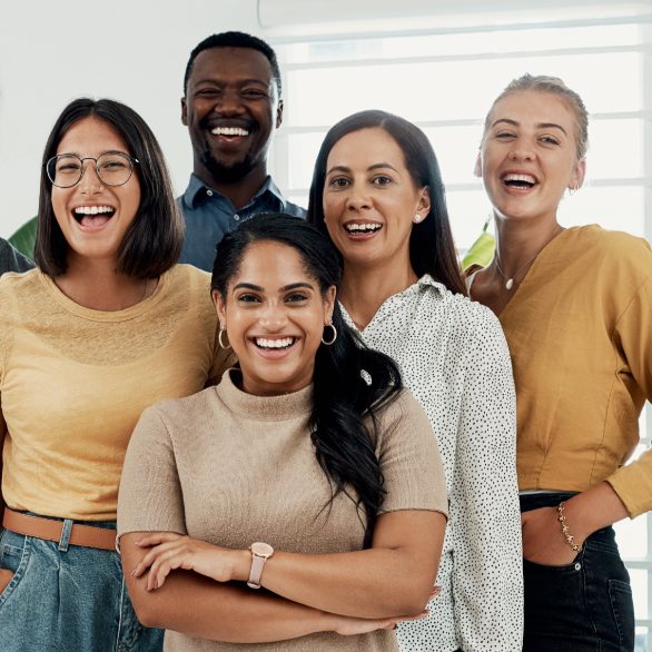 Five people stand close together indoors, smiling at the camera, dressed in casual and business-casual attire, with natural light in the background.