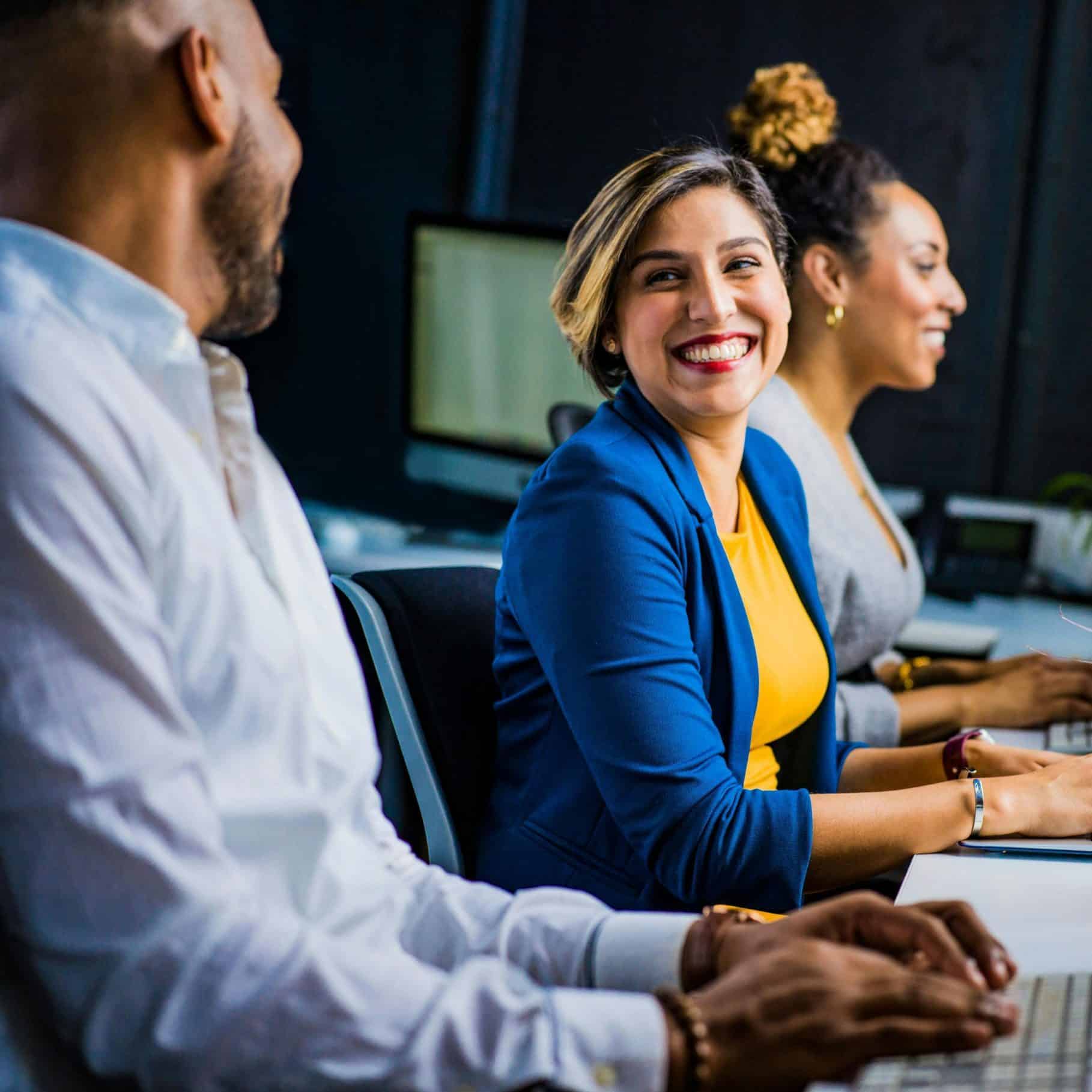 Three people sit at desks with computers in an office setting. The woman in the center smiles brightly at a colleague.
