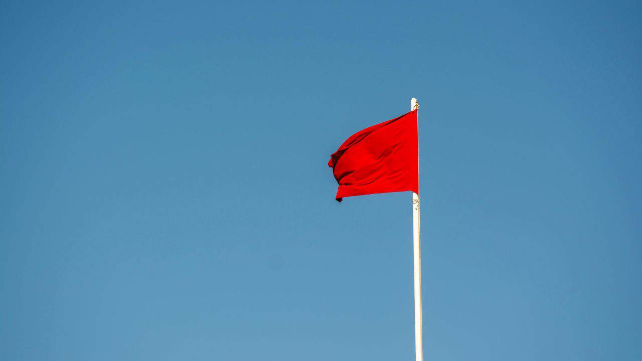 A plain red flag is flying on a white flagpole against a clear blue sky.