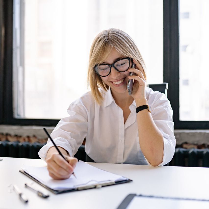 pexels-yankrukov-7793187 (1) Women with short blond hair and dark framed glasses writing notes at a desk while on the phone
